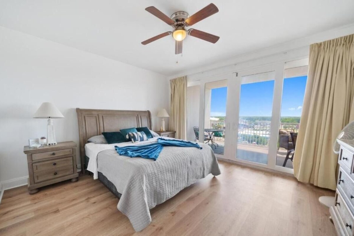 A bedroom with a bed, nightstands, a ceiling fan, and a view through large windows.