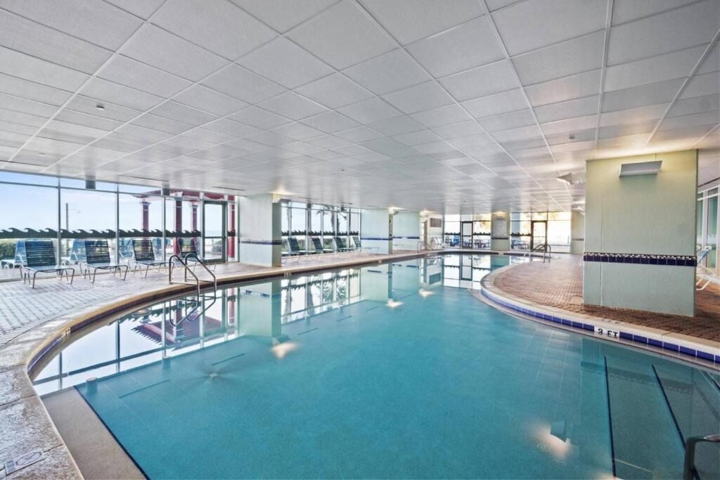 Una piscina interior con paredes de vidrio transparente, piso de baldosas y algunas sillas de descanso.