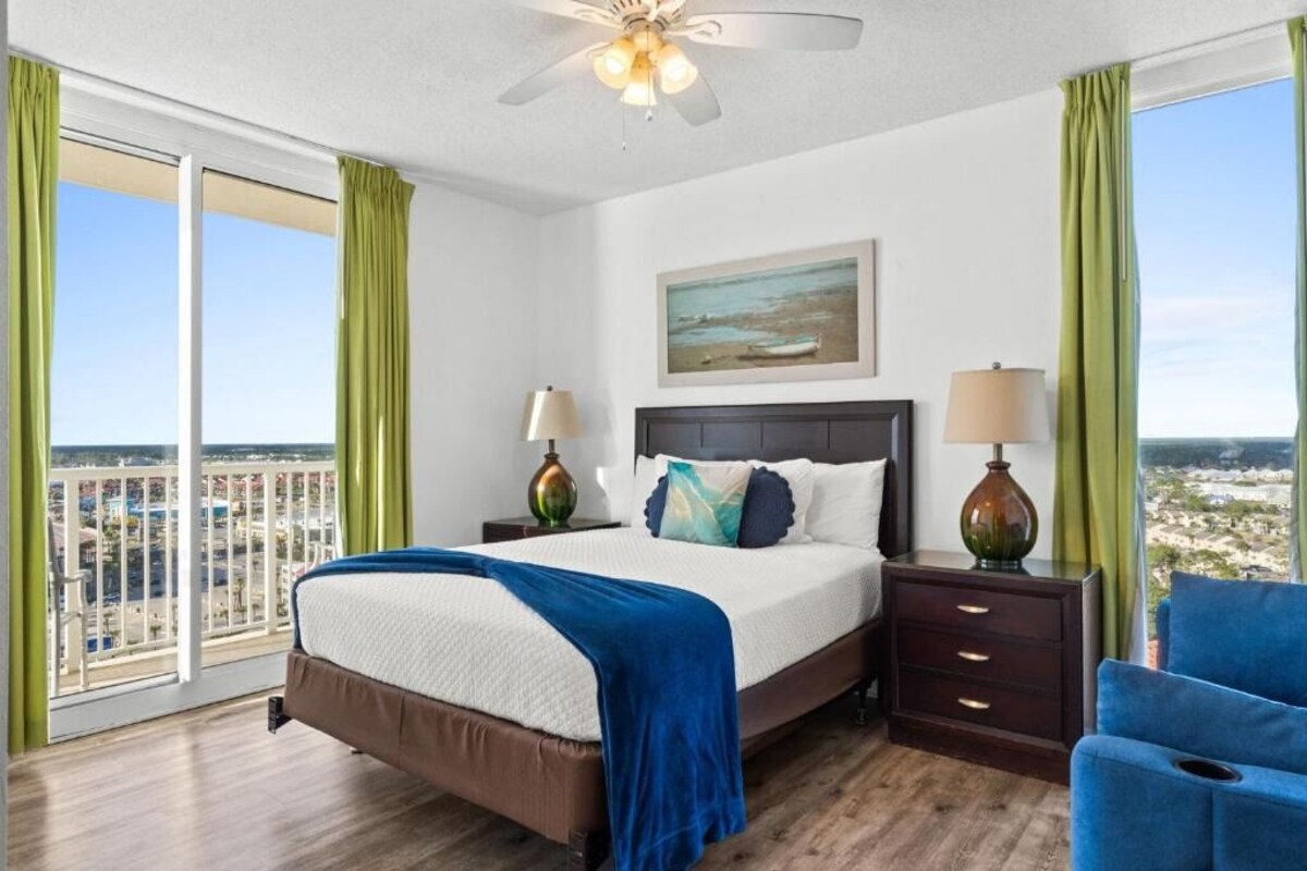 A hotel room with a bed, two bedside tables, a ceiling fan, a painting, and a view of the city.