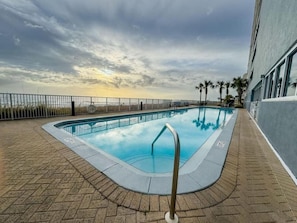 A swimming pool with a metal ladder, surrounded by a brick deck and a fence, with palm trees and a building in the background. - Ocean Reef (Panama City Beach)