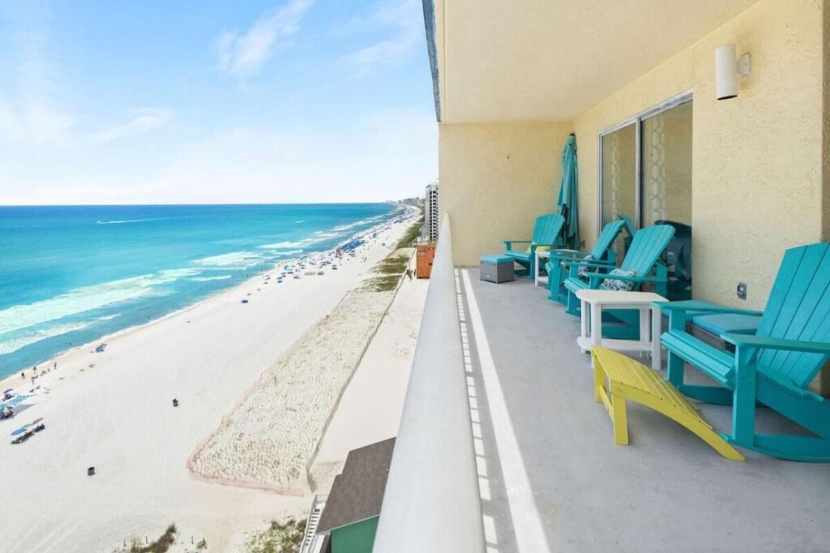 A balcony with turquoise chairs and a yellow Adirondack chair overlooking a beach with a sandy shore and ocean.