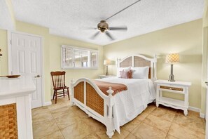 A bedroom with a white bed, a ceiling fan, a window with blinds, a chair, and a nightstand with a lamp. - Regency Towers (Panama City Beach)