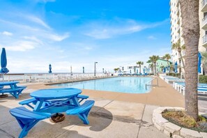 A poolside area with blue picnic tables and umbrellas, a clear sky, and a building with balconies in the background. - Regency Towers (Panama City Beach)