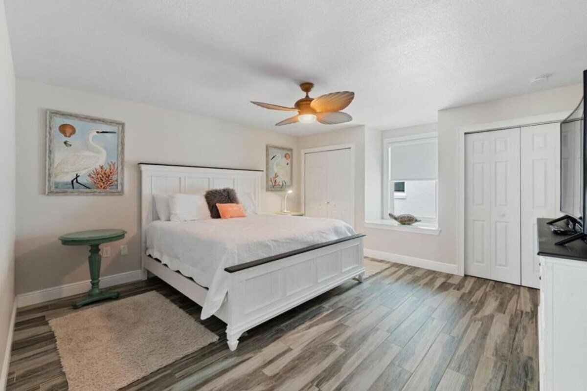 A bedroom with a bed, a ceiling fan, a green side table, and artwork on the walls.