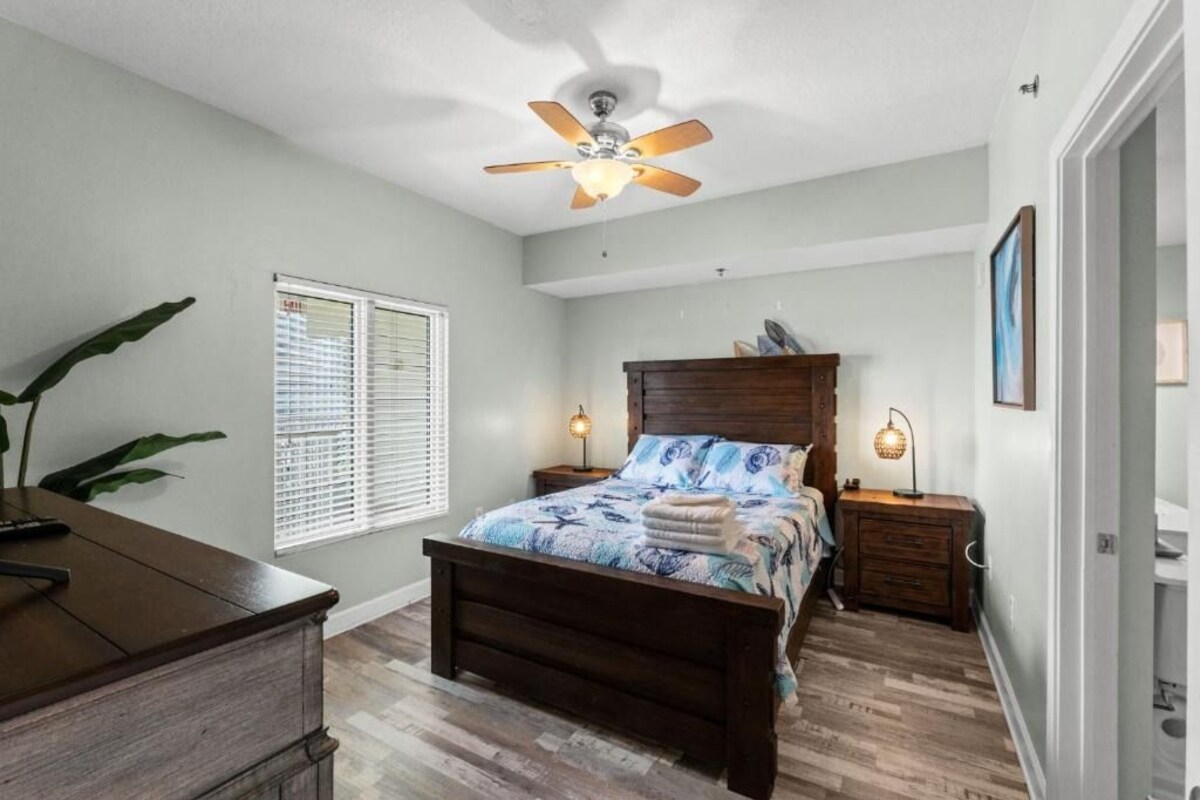 A bedroom with a wooden bed, a ceiling fan, bedside lamps, a nightstand, a window with blinds, and a plant.