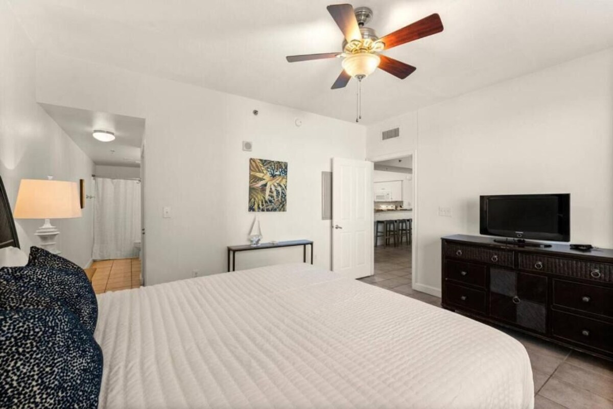 A hotel room with a bed, a ceiling fan, a television, a dresser, and a painting on the wall.