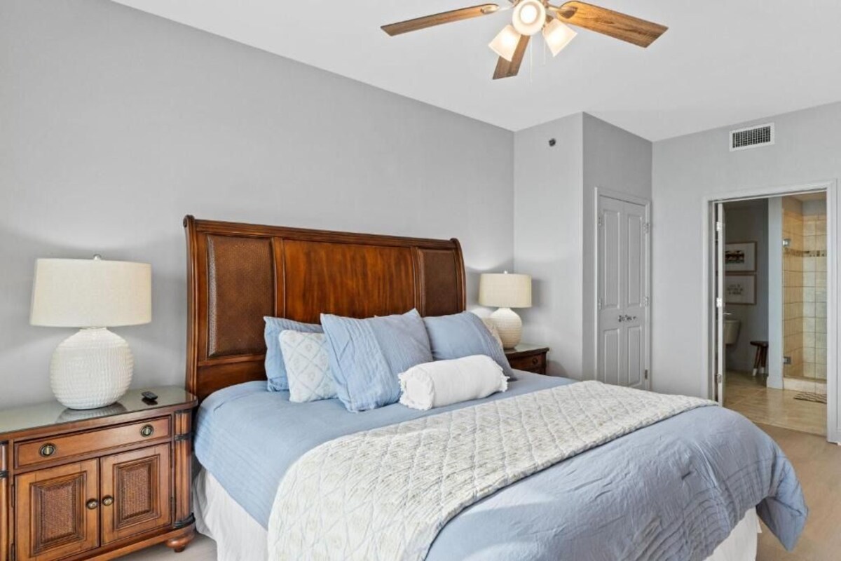 A bedroom with a wooden headboard, two bedside lamps, a bed with blue bedding, and a ceiling fan.
