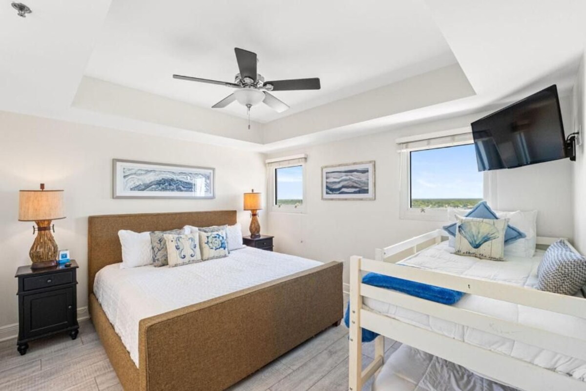 A bedroom with a bed, a ceiling fan, a TV mounted on the wall, and framed artwork on the walls.