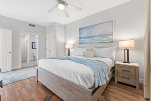 A bedroom with a bed, two bedside tables, a ceiling fan, a painting, and a door leading to another room. - Sterling Breeze (Panama City Beach)