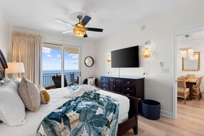 A hotel room with a bed, a ceiling fan, a TV, a dining area with a table and chairs, and a view of the ocean. - Sterling Breeze (Panama City Beach)