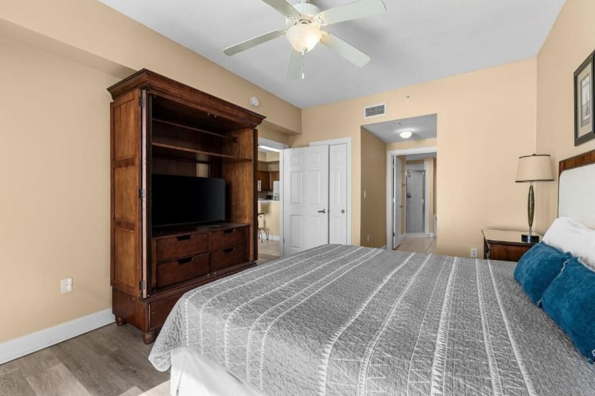 A bedroom with a large bed, a wooden cabinet, a ceiling fan, and a door leading to another room.