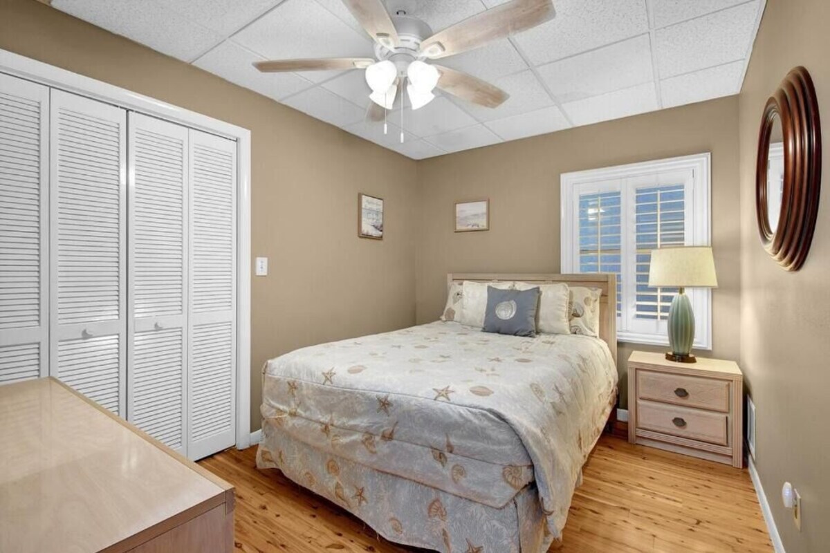 Ein Schlafzimmer mit Bett, Deckenventilator, Fenster mit Jalousien, Nachttisch mit Lampe und Kleiderschrank mit Klappläden.