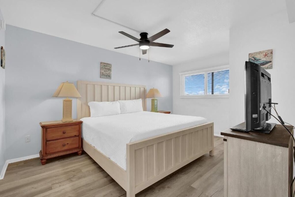 A bedroom with a bed, nightstands, a ceiling fan, a window with blinds, and a TV on a stand.