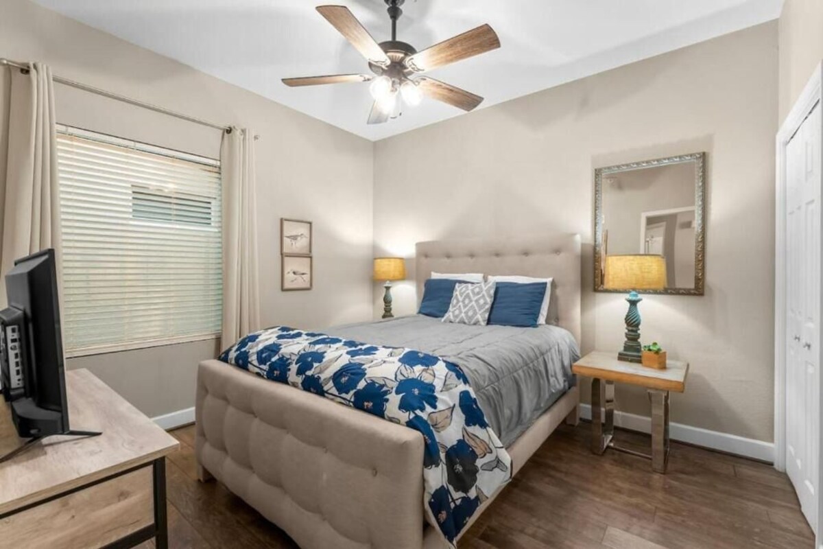 A bedroom with a bed, bedside lamps, a ceiling fan, a TV, and a window with blinds.