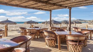 Un ristorante sulla spiaggia con tavoli e sedie in legno, ombrelloni di paglia e vista sull'oceano.