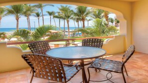 Een balkon met een ronde tafel en vier stoelen, met uitzicht op een strand met palmbomen en de oceaan.
