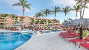 Zwembadgebied van een resort met helder uitzicht op het zwembad, ligbedden, palmbomen en een gebouw met balkons.