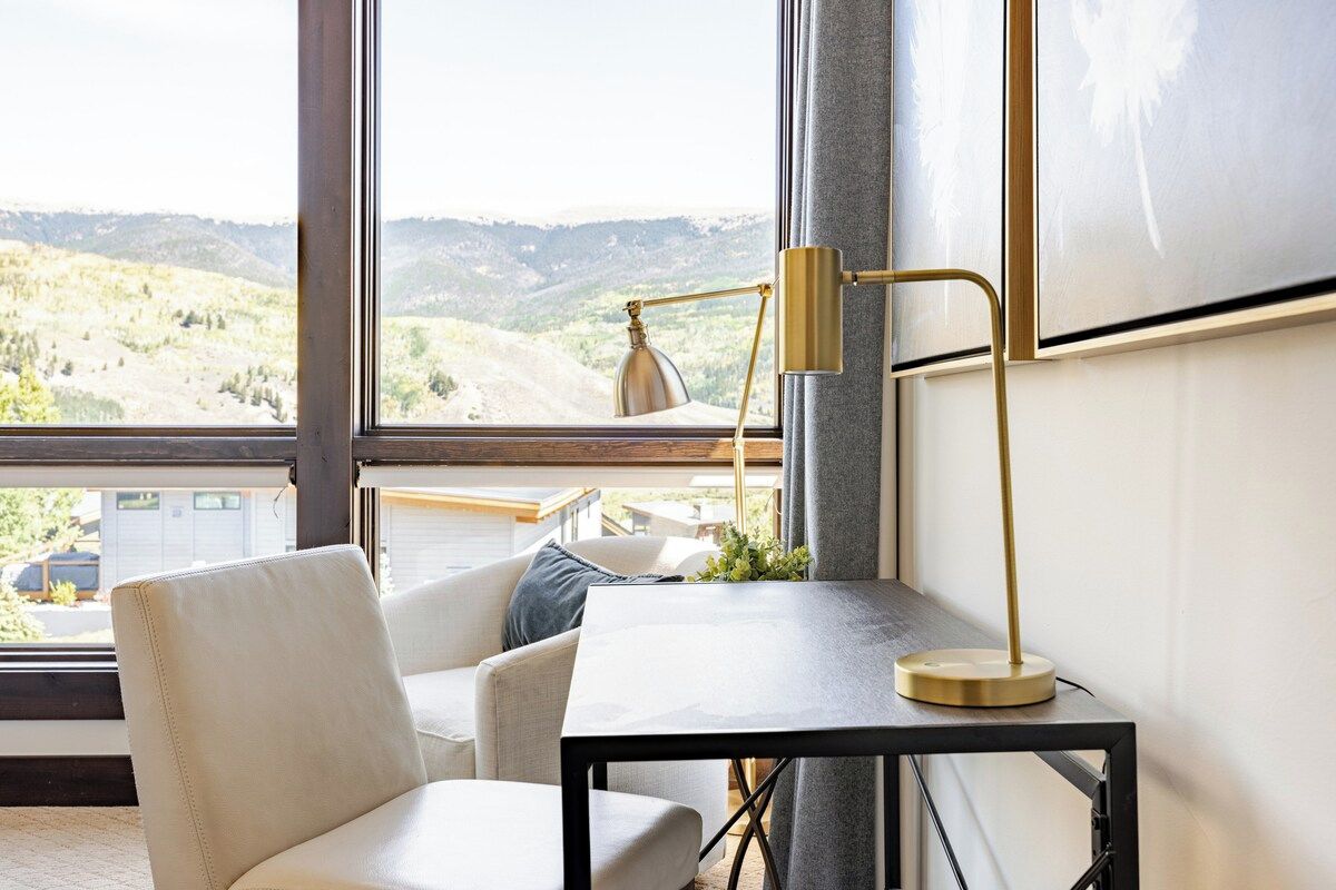 A modern room with a view of a mountainous landscape through large windows, a white leather chair, a small table, and a floor lamp.