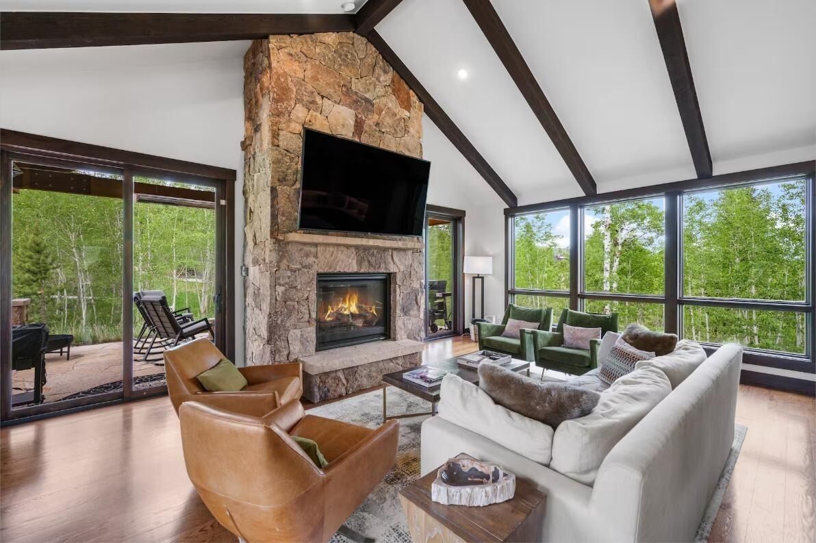 A spacious living room with a stone fireplace, comfortable seating, and large windows offering a view of the outdoors.
