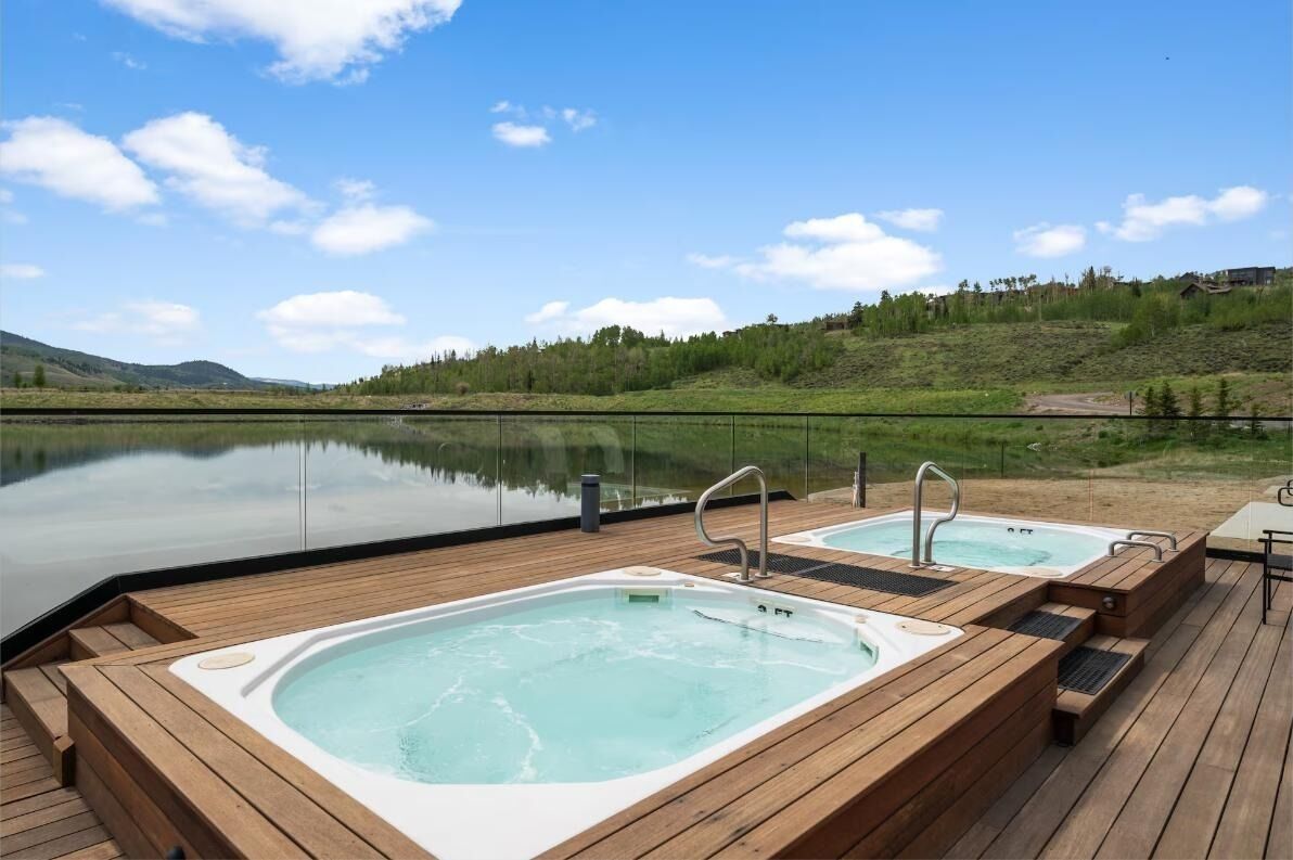 A wooden deck with two hot tubs overlooking a calm lake and a hilly landscape.