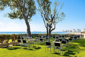 An outdoor seating area with tables and chairs arranged on grass, overlooking a beach and city skyline.