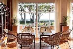 A room with wicker chairs and a table, large windows, and a view of the outdoors.