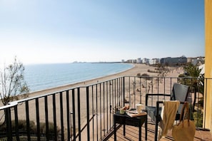 A balcony with a view of the beach and ocean, a table with a chair, and a bag.