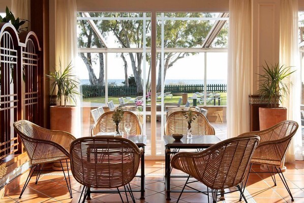 A room with wicker chairs and a table, large windows, and a view of the outdoors.