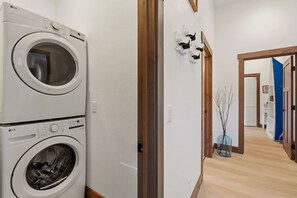 Une buanderie équipée de deux machines à laver superposées, d’un cadre de porte en bois, d’un luminaire fixé au mur et d’un couloir menant à une autre pièce.