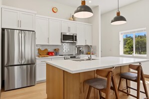 Une cuisine moderne dotée d’appareils électroménagers en acier inoxydable, d’un grand îlot central avec des tabourets, et offrant une vue sur l’extérieur grâce à une fenêtre.