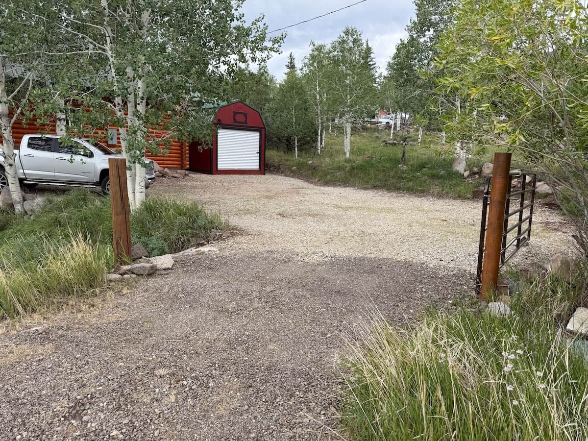Une allée de gravier mène à une grange rouge dont la porte du garage est fermée, le tout étant entouré d’arbres et d’un véhicule garé.