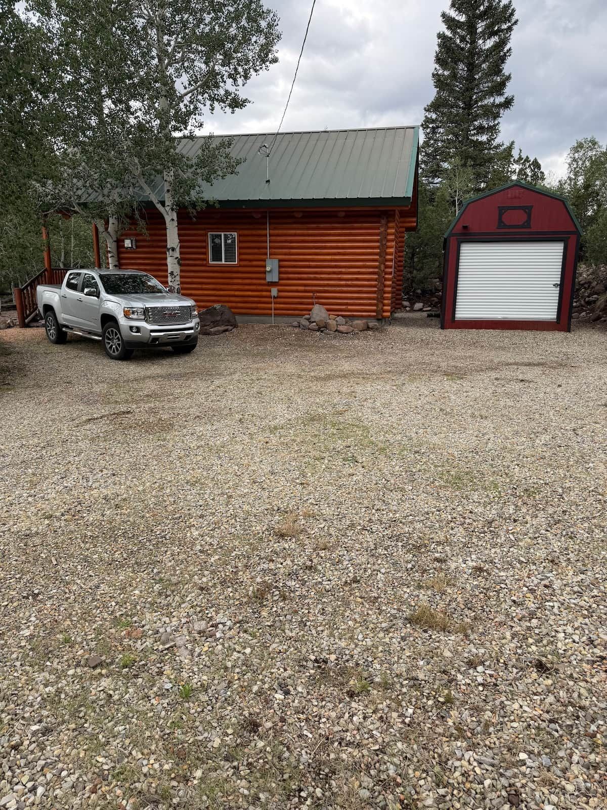 A cabin with a gravel driveway, a white pickup truck parked in front, and a red barn to the right.