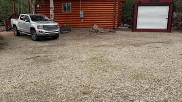A cabin with a gravel driveway, a white pickup truck parked in front, and a red barn to the right.