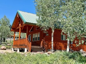 A log cabin with a wooden porch and a metal roof, surrounded by trees and rocks. - Skoots Creek Hideaway — a cozy, classic log cabin! (Panguitch Lake)