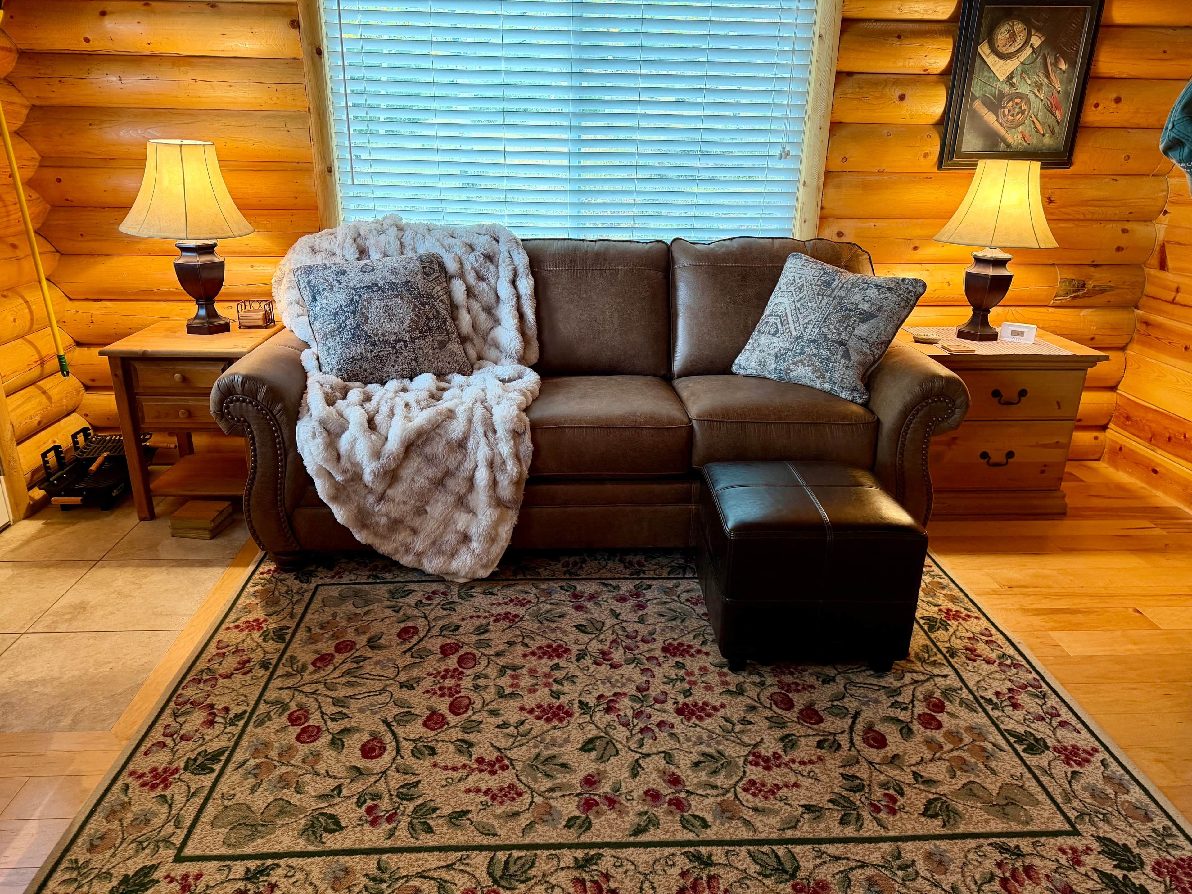 A cozy log cabin interior with a brown leather sofa, a matching ottoman, and a floral area rug. There are two wooden nightstands with lamps and a framed picture on the wall.