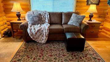 A cozy log cabin interior with a brown leather sofa, a matching ottoman, and a floral area rug. There are two wooden nightstands with lamps and a framed picture on the wall.