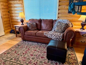 A cozy room with a brown leather sofa, patterned throw pillows, a black ottoman, and a floral area rug. - Skoots Creek Hideaway — a cozy, classic log cabin! (Panguitch Lake)