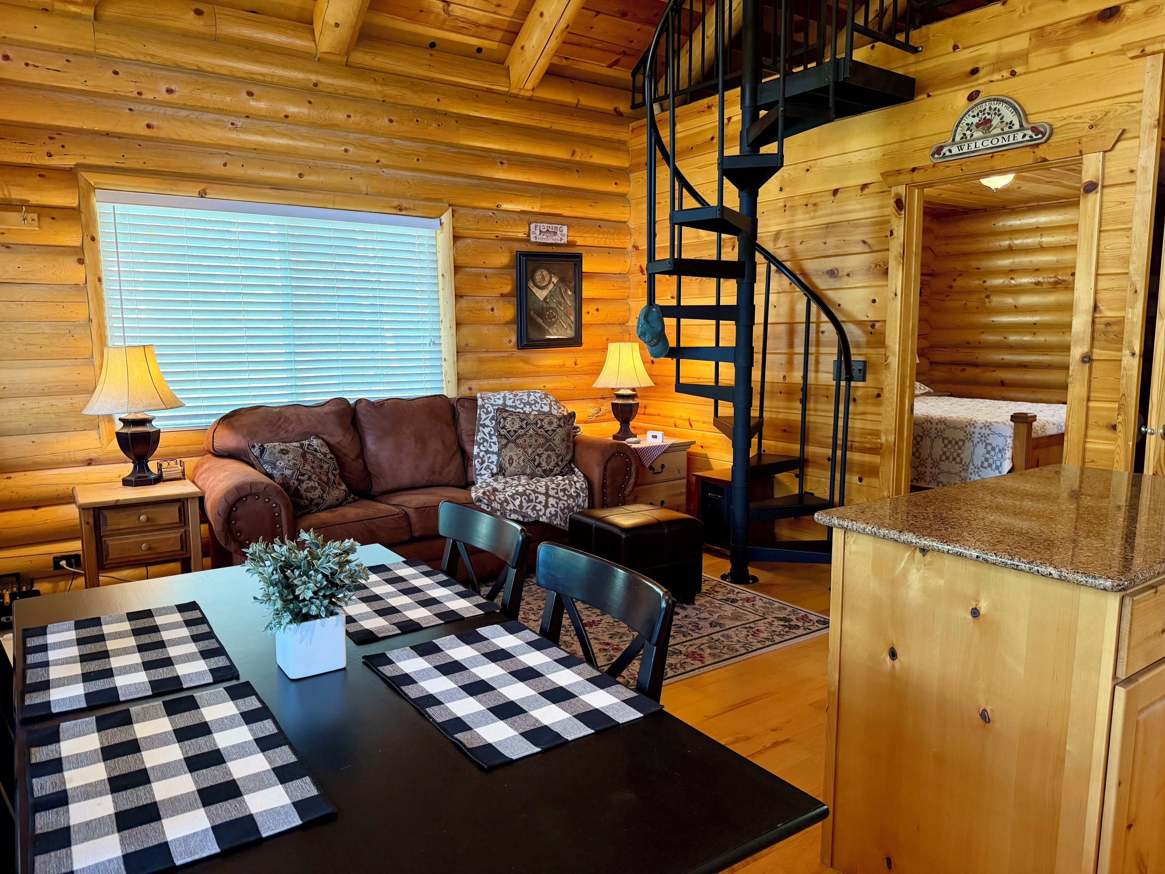 A cozy cabin interior with a wooden interior, a sofa, a dining table with checkered placemats, and a spiral staircase.