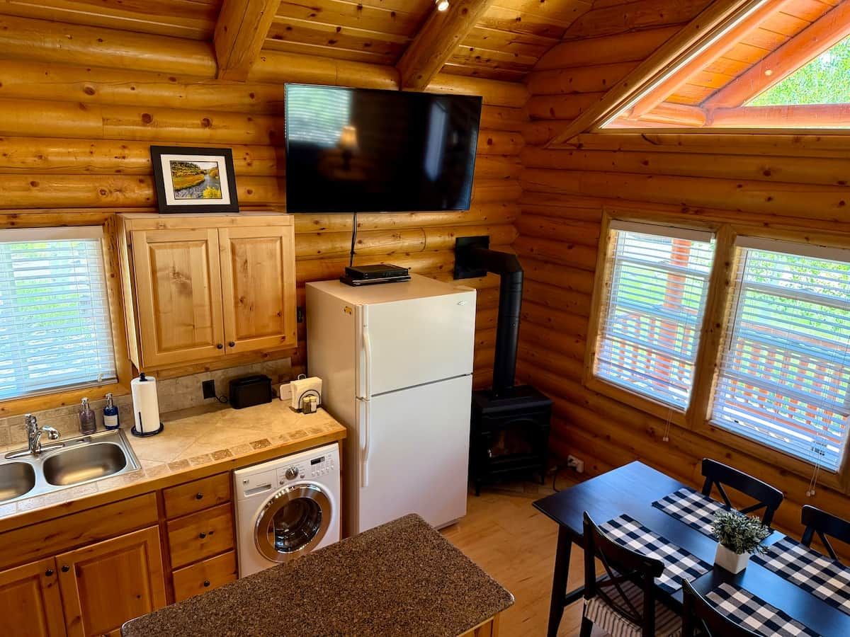 Interior de una cabaña de troncos con cocina, lavadora, mesa de comedor y televisor montado en la pared.
