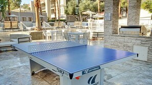 Ein Tischtennistisch im Freien, mit einer Steinwand im Hintergrund, Sitzgelegenheiten und einem Schild für Tischtennisartikel.