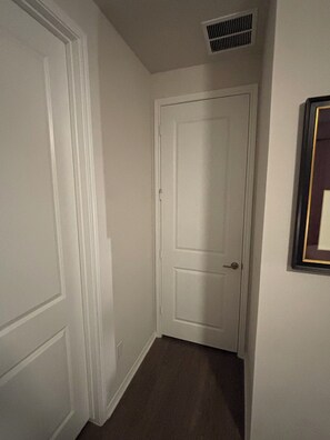Ein Flur mit einer weißen Tür, einem Lüftungsgitter an der Decke und einem gerahmten Bild an der Wand.