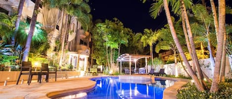 A nighttime swimming pool area with a curved design, surrounded by palm trees and outdoor seating.
