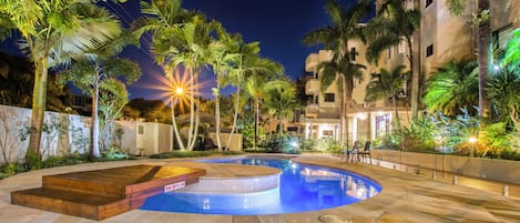 A nighttime pool area with a tiled deck, palm trees, and a building with balconies.