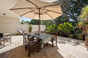 Patio esterno con tavolo e sedie, circondato da piante in vaso e un ombrellone.