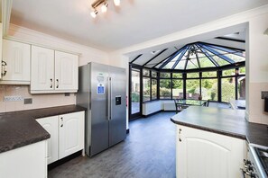 A modern kitchen with white cabinetry, a stainless steel refrigerator, and a large windowed area leading to a conservatory.