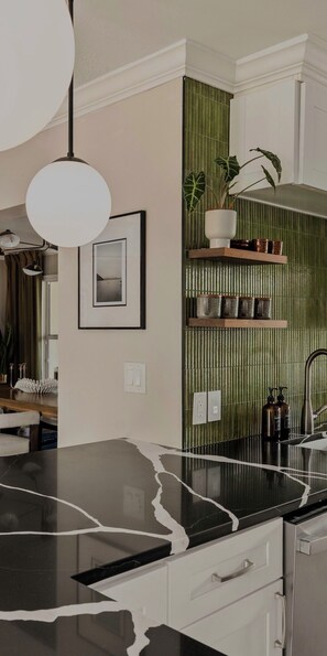 Una cocina moderna con encimera de mármol negro, armarios blancos, una lámpara colgante, un cuadro en la pared y un panel vertical con jardín.