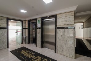 Eine Hotellobby mit einer Rezeption, zwei Aufzügen und einem schwarzen Teppich mit goldener Schrift.