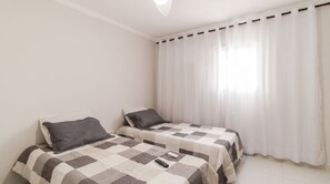 Ein Schlafzimmer mit zwei Betten, jeweils mit einem gemusterten Überwurf, einer weißen Wand und einem Fenster mit durchscheinenden Vorhängen.