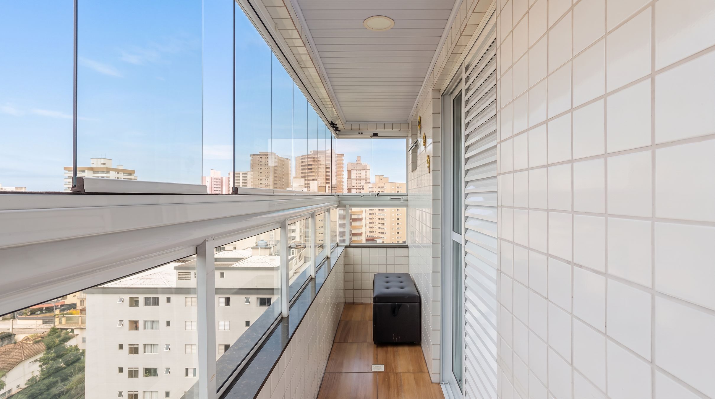 Un balcón con vista a la ciudad, paredes de baldosas blancas y un caja de almacenamiento negra.
