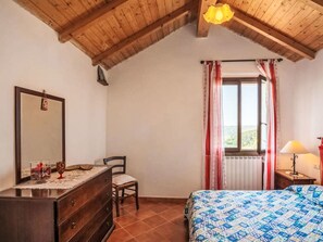 A bedroom with a wooden ceiling, a bed with a patterned bedspread, a dresser, a chair, a window with curtains, and a nightstand with a lamp. - Viletta Ulivo by Interhome (Diano Marina)